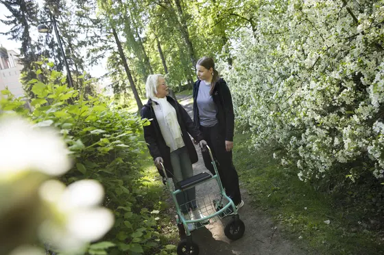 Two people standing in a garden. The other one has got wheeled walker.