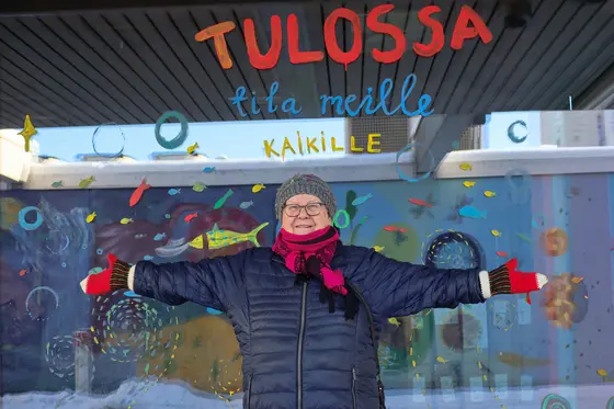 Liisa Aunula kädet sivuille avoinna värikkään ikkunan edessä.