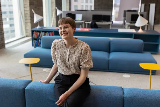 Landscape architect Sofia Kangas smiles as she leans back on a sofa in the large office room in her workplace, which is filled with modern furniture and lighting. 