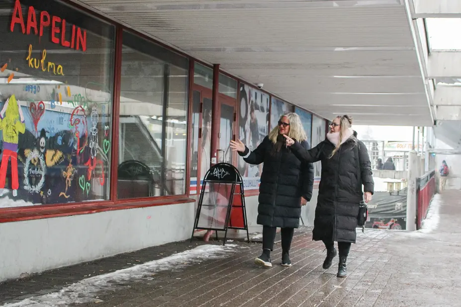 Two women wearing black padded jackets are walking in front of a colorful window.