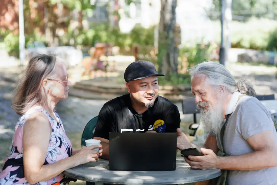 Three people sitting at a table outside and looking at a laptop and a phone.