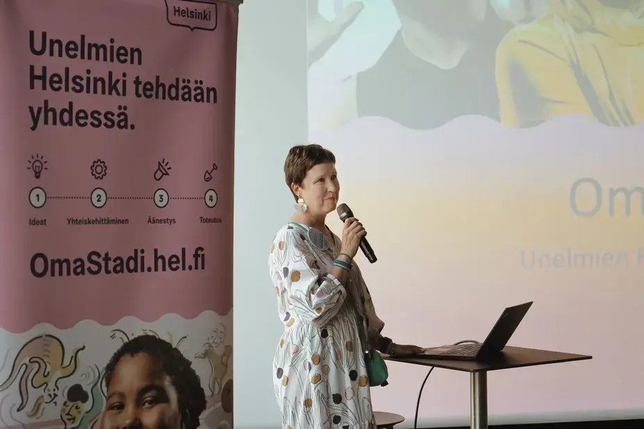 Kirsi Verkka speaks into the microphone in front of the screen.