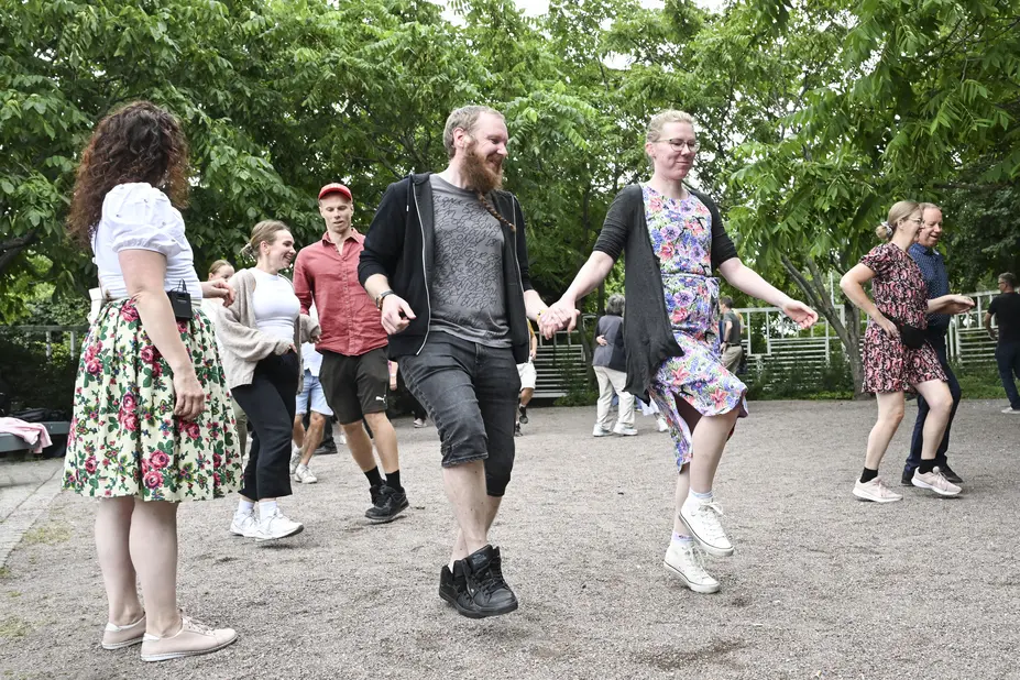 Smiling people are dancing in pairs in the leafy courtyard of Malmitalo. A woman is standing there watching them.
