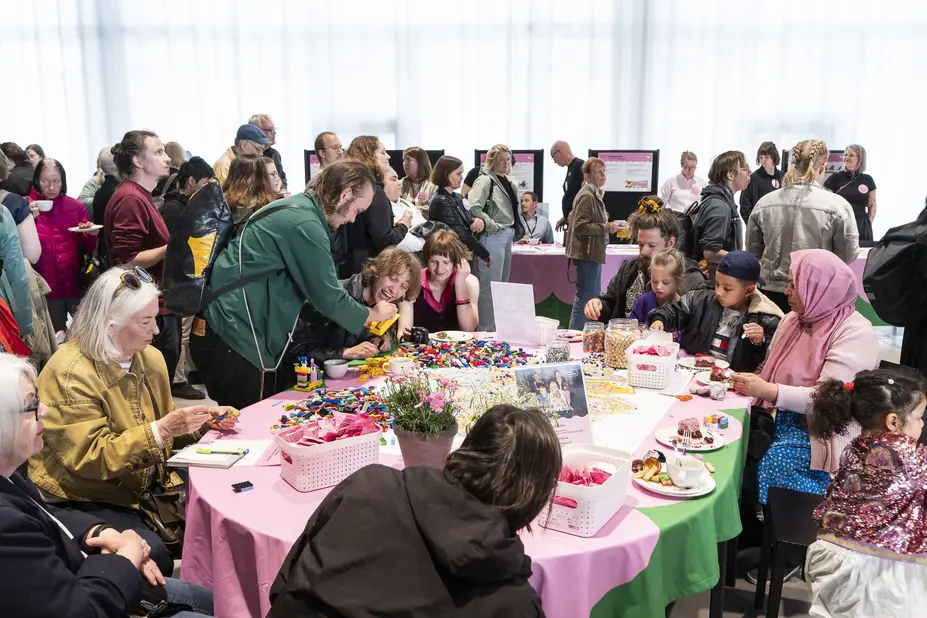 A large number of city residents of different ages and from different cultural backgrounds are gathered at Oodi for the OmaStadi co-creation event. Some of them are sitting at a table and others are standing.