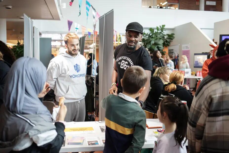 Two people behind a table smiling and talking to three children on the other side of the table.  