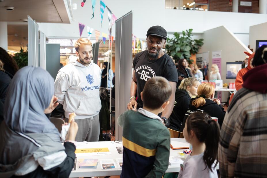 Two people behind a table smiling and talking to three children on the other side of the table.  