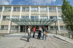 Opiskelijat osoittavat iloisina kohti Alppilan lukion nimikylttiä koulun ovien yläpuolella,