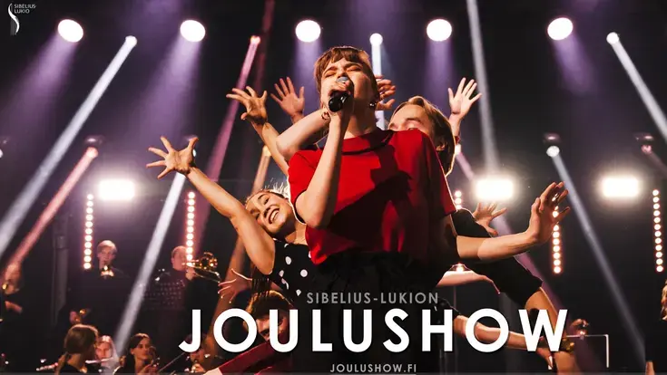 Joulushown laulaja punaisessa asussa etualalla ja tanssijoita tanssijoita kädet ylhäällä.