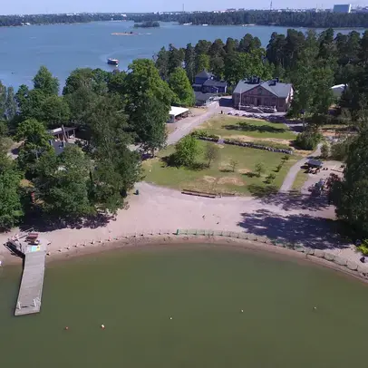 Saari ilmakuvassa, hiekkaranta, laituri, nurmialue ja rakennus.