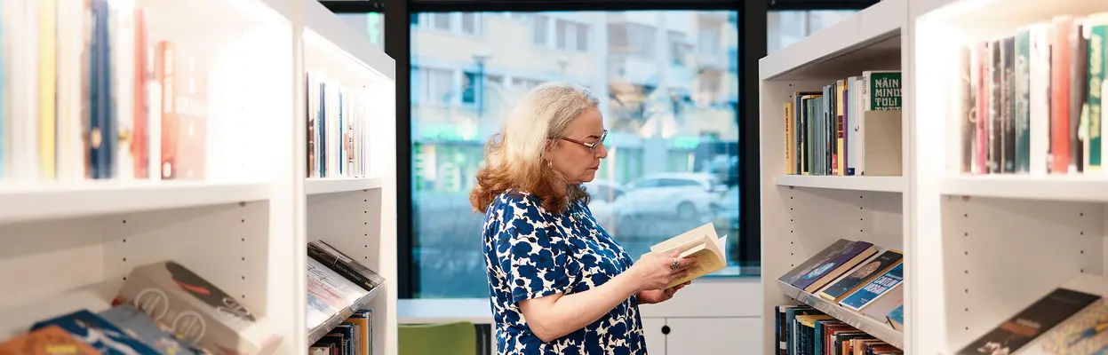 Nainen lukee kirjaa Maunulan kirjaston kirjahyllyjen välissä tämä ruotsiksi