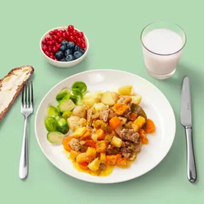 : A plate with chopped Brussels sprouts, wedges of new potatoes, and a small portion of a casserole made with roasted root vegetables and broad beans, served with rye bread, a glass of milk, and a small bowl of berries.
